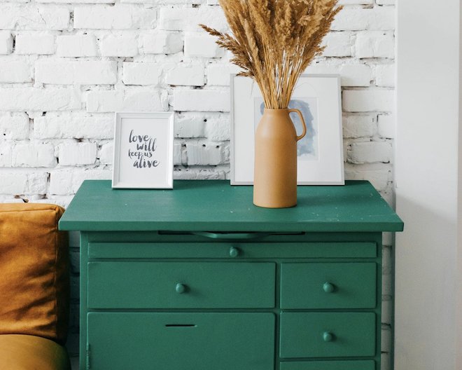 Green dresser with four drawers a vase of dried flowers and and a white portrait with the word love will keep us alive written in black ink.